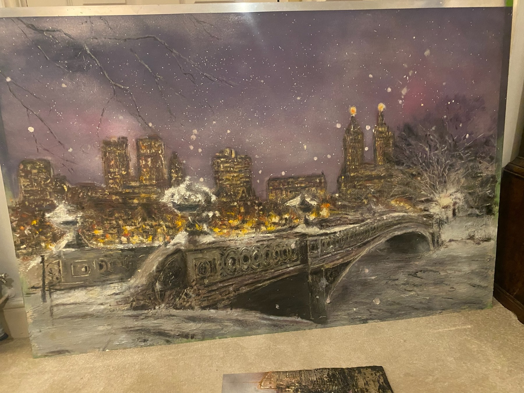 Bow Bridge in Central Park beneath a purple sky, covered in snow with golden city lights