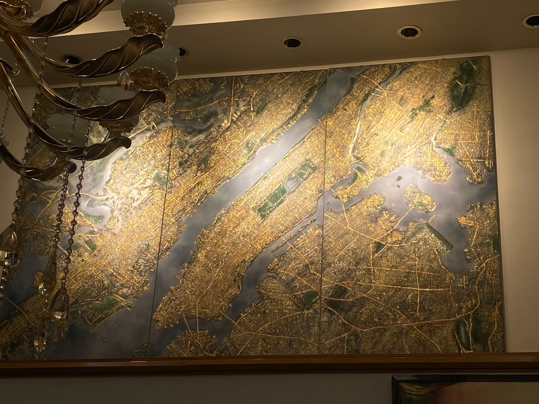 A large three-panel gilded artwork depicting New York and its waterways, installed beneath a chandelier